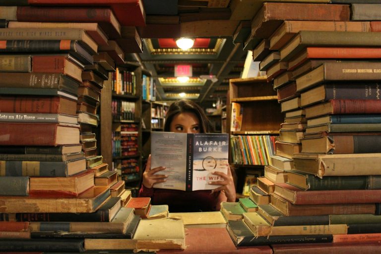 Vrouw leest een boek omringd door stapels boeken in een bibliotheek.