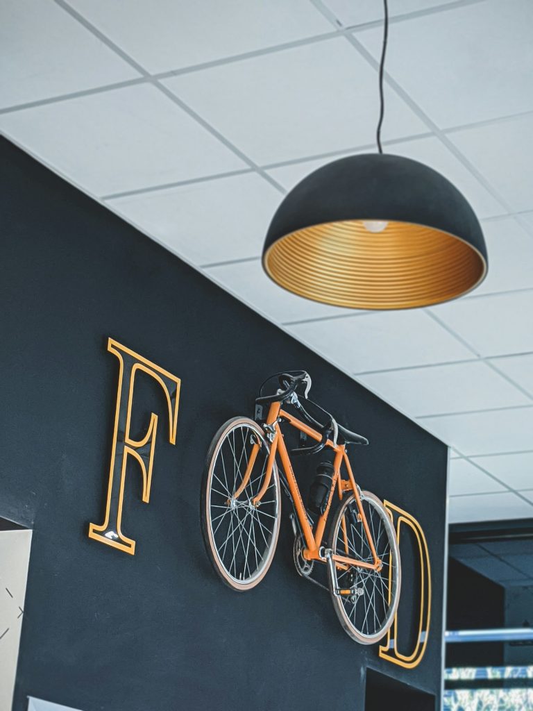 Zwarte wand met een oranje fiets en metalen letters die "F" en "D" vormen.