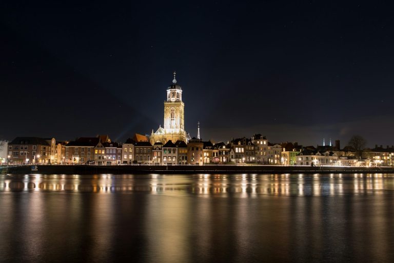 Nachtzicht op de stad Deventer met een toren, verlicht door straatlampen en reflecties in het water.