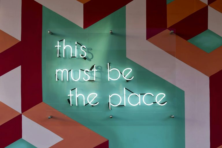 Neon tekst "this must be the place" op een kleurrijke geometrische achtergrond.