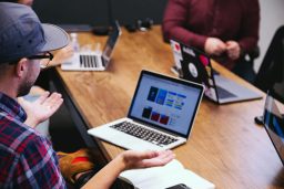 Man met bril en pet in vergadering, kijkt naar laptop met kleurrijke ontwerpen.